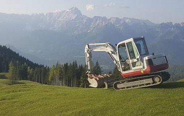 Auf dem Bild ist eine grüne Wiese im Vordergrund und eine blaue Alpenlandschaft im Hintergrund zu sehen, rechts mittig steht ein weißer Bagger.