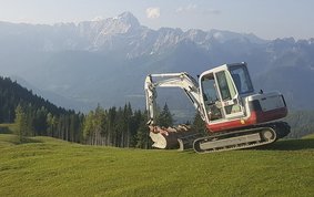 Auf dem Bild ist eine grüne Wiese im Vordergrund und eine blaue Alpenlandschaft im Hintergrund zu sehen, rechts mittig steht ein weißer Bagger.