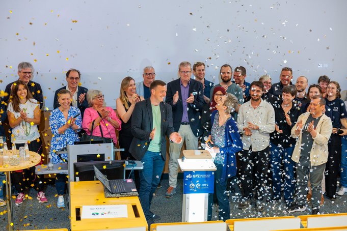 A group of about 20 people is standing in front of a white wall, applauding and enjoying the confetti rain down on them.