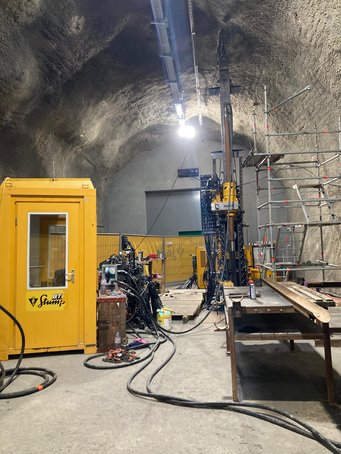 Eine ca. 10 Meter hohe Tiefbohranlage steht in einem Untertage-Tunnel, daneben ein Gerüst, Leitungen und weiteres Gerät sowie eine gelbe, telefonzellengroße Steuerkabine.