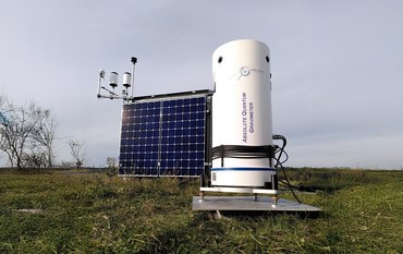 On a green field stands a white, barrel-shaped container with an inscription, in which the sensitive measuring device is installed. Next to it is a solar panel.