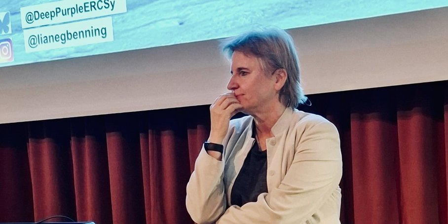 Eine Frau steht unten gegen eine schwarze Bühne gelehnt und hört einer Person aus dem Publikum zu.