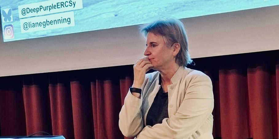 A woman stands leaning against a black stage and listens to a person from the audience.