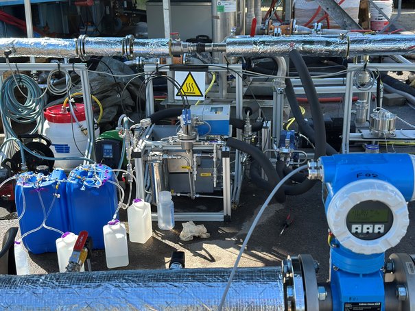 Nahaufnahme einer technischen Anlage mit Kanistern, Schläuchen, Rohren und Pumpen.