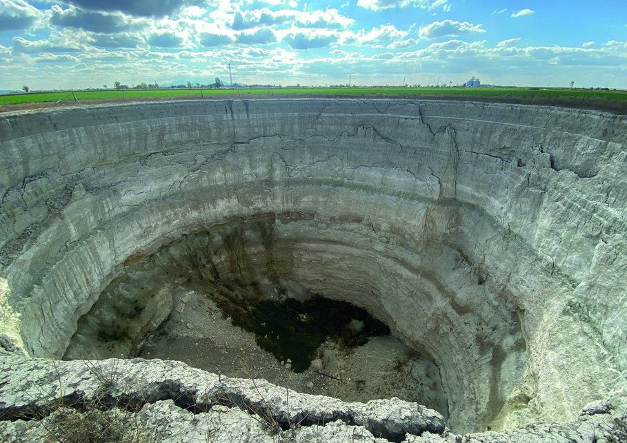 The photo shows a deep sinkhole in the karst terrain, surrounded by green landscape and blue sky.