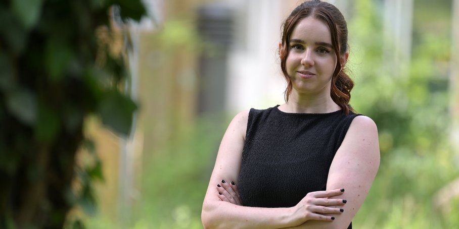 [Translate to English:] Portrait einer jungen Frau mit langem Haar und schwarzem Kleid vor einem verschwommenen grünen Park-Hintergrund
