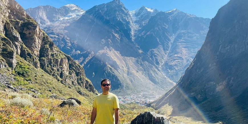 A young man with dark short hair and sunglasses, wearing a yellow T-shirt, stands in the mountains. In the background is a valley with houses standing close together.