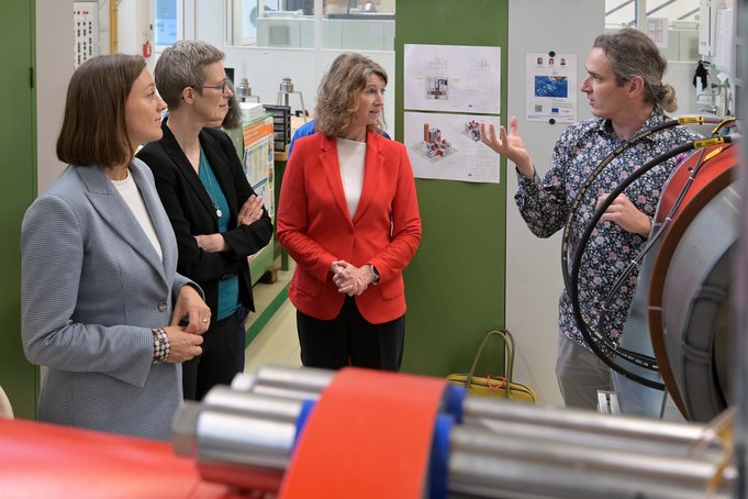In einem großen Labor steht ein Mann an einer großen roten Maschine und erläutert die Funktionsweise. Drei Frauen stehen daneben und sehen interessiert zu.