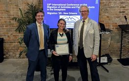 Foto von der Preisträgerin mit den Hauptorganisatoren vor einem Bildschirm auf dem der Titel der Konferenz steht