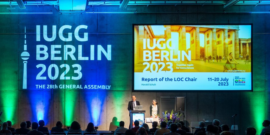 Two persons standing on stage and speaking, with the IUGG logo superimposed over her on a large screen and projected on the wall. The room is dark and many people are seated as spectators.
