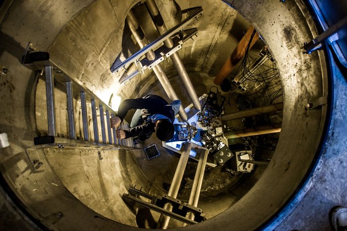 In a round, concrete chamber resembling a well, 15 metres below ground, a person climbs down a ladder. At the bottom is a triangular structure, on each side connected to the wall by two pipes.