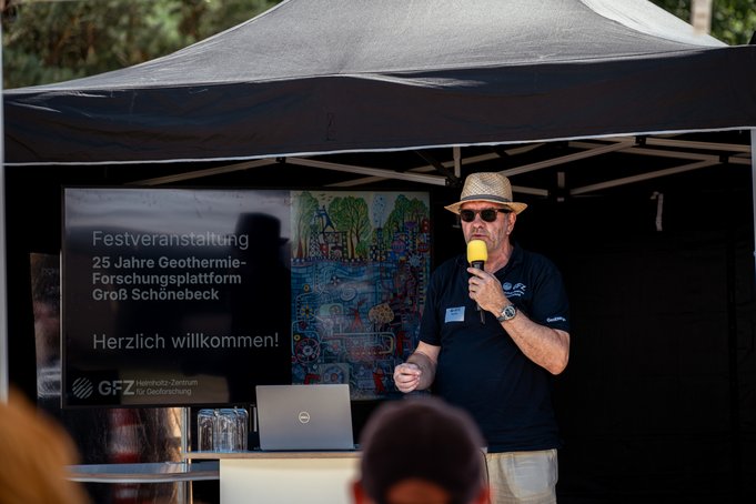 Ein Mann mit Hut und Sonnenbrille steht unter einem Zeltdach und hält ein Mikrofon. Daneben ein Bildschirm.