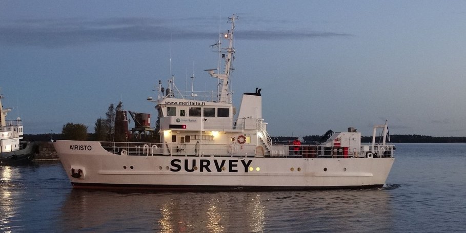 Das Foto zeigt das finnische Vermessungsschiff „Airisto“ etwa 100 Meter vom Betrachter entfernt im Hafenbecken des finnischen Ortes Mäntyluoto.  Es hat gerade abgelegt und ist im Begriff, den Hafen zu verlassen. Das Schiff hat dem Betrachter die Backbord-Seite zugewandt. Es ist etwa 30 Meter lang und 15 Meter hoch und trägt auf der Seite in Großbuchstaben die Aufschrift „SURVEY“. Oben über der Brücke steht die Aufschrift www.meritaito.fi. Das ist die Webseite der Reederei dieses Schiffes.