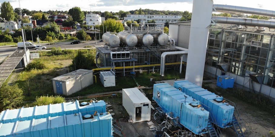 Der Bohrplatz: Im Vordergrund vier blaue und drei Weiße Container mit Treppen und Rohren. Dahinter eine größere Anlage mit dicken Rohren und Flüssigkeitstanks.