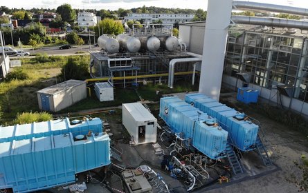 Der Bohrplatz: Im Vordergrund vier blaue und drei Weiße Container mit Treppen und Rohren. Dahinter eine größere Anlage mit dicken Rohren und Flüssigkeitstanks.