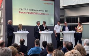 Publikum und 6 Diskutierende mit einer Moderatorin auf dem Podium (stehend)