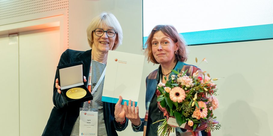 Zwei Frauen stehen nebeneinander. Die Rechte hat eine Medaille und eine Urkunde in der Hand, die linke einen Blumenstrauß. Beide schauen freundlich in die Kamera.