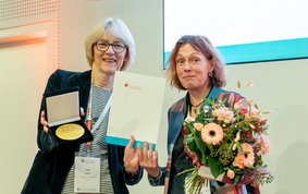 Two women standing side by side. The woman on the right is holding a medal and a certificate, while the woman on the left is holding a bouquet of flowers. Both are smiling at the camera.