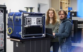 Ein blauer Kasten mit einem Messgerät auf einem Rollkontainer. Daneben stehen zwei Personen und blicken freundlich in die Kamera.