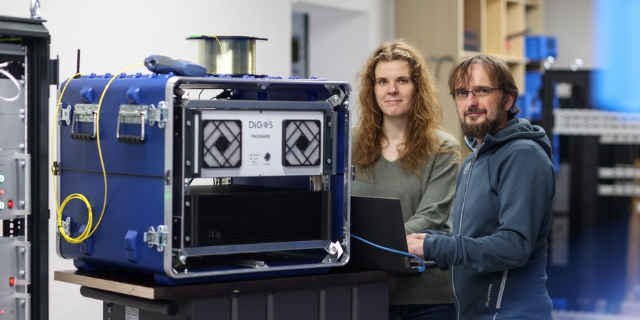 Ein blauer Kasten mit einem Messgerät auf einem Rollkontainer. Daneben stehen zwei Personen und blicken freundlich in die Kamera.