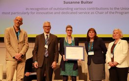 5 Personen auf der EGU vor einem Bildschirm. Frau Buiter hält den Preis (Urkunde in Rahmen)