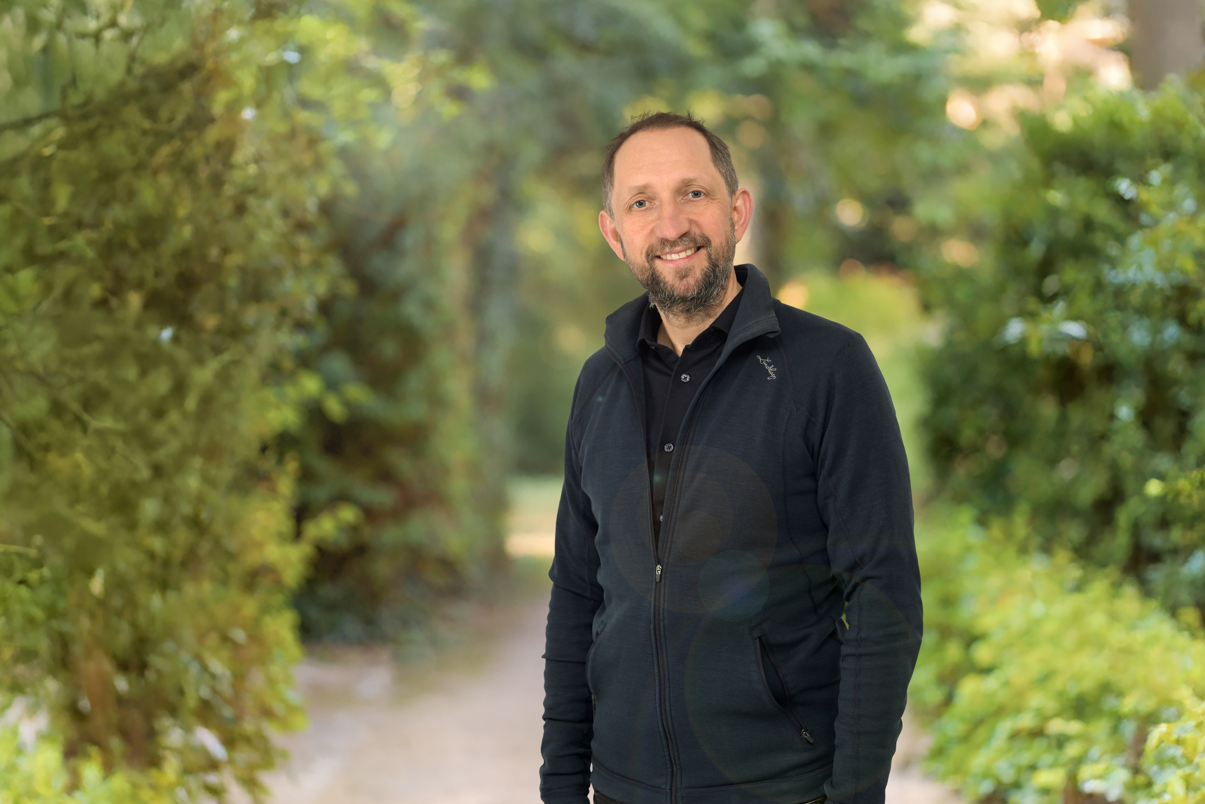 Portrait of a man in front of blurred green bushes: The man is wearing dark clothes, has a beard and is smiling warmly.