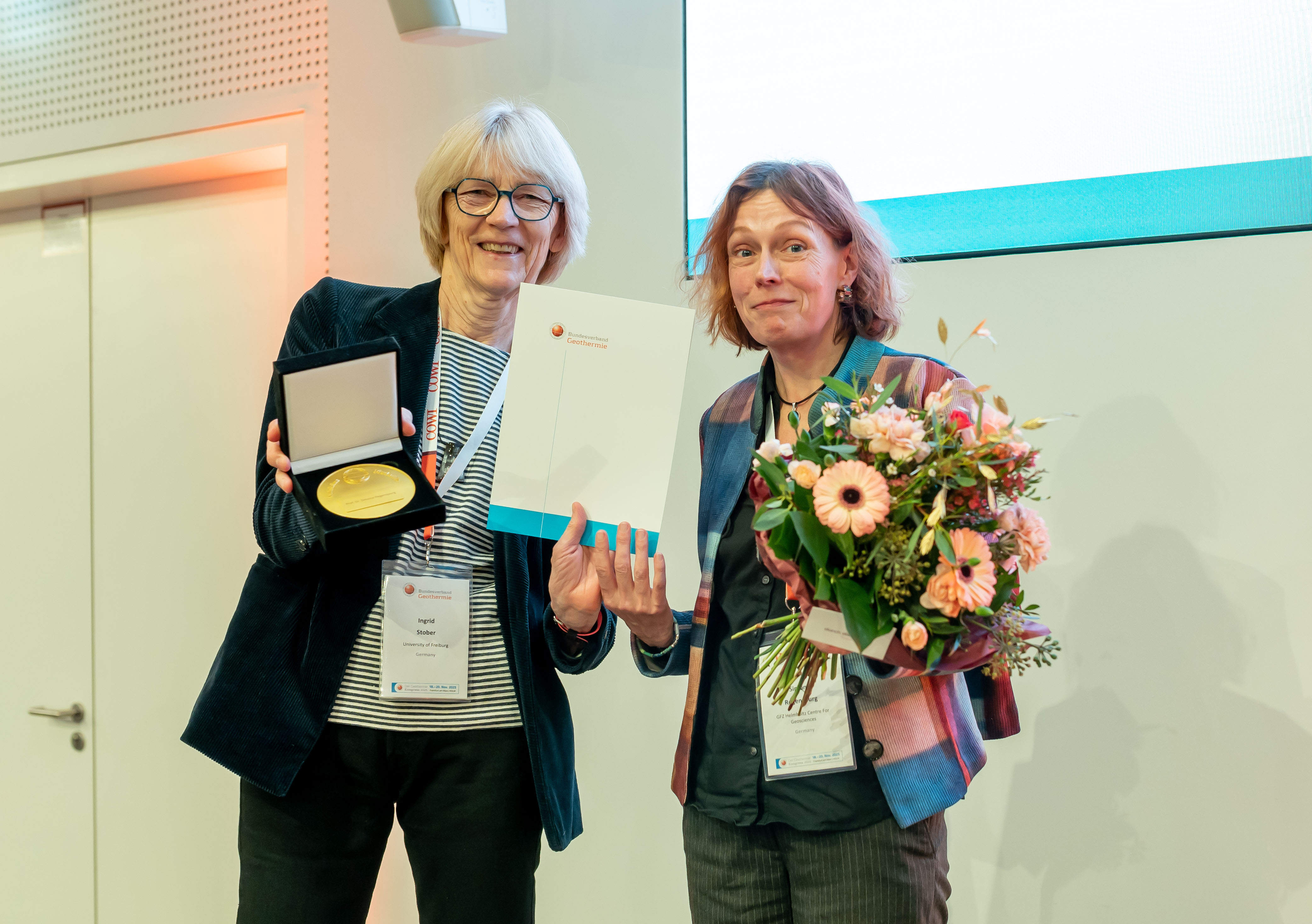 Two women standing side by side. The woman on the right is holding a medal and a certificate, while the woman on the left is holding a bouquet of flowers. Both are smiling at the camera.