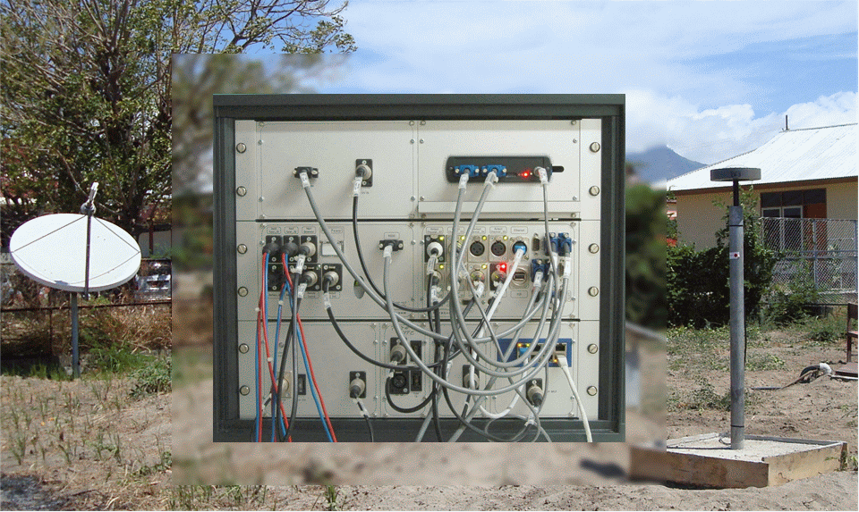 Figure, center: Front-side of a GITEWS GPS RTR station with modules and cables; background: VSAT-antenna for satellite communication (left) and GPS-antenna (right), both on concrete fundaments. The graphic shows a collage. In the middle it shows the front-side of a GITEWS GPS RTR station with several modules and cables. In the background a VSAT-antenna for satellite communication is visible on the left side and a GPS-antenna on the right side, both mounted on concrete fundaments.